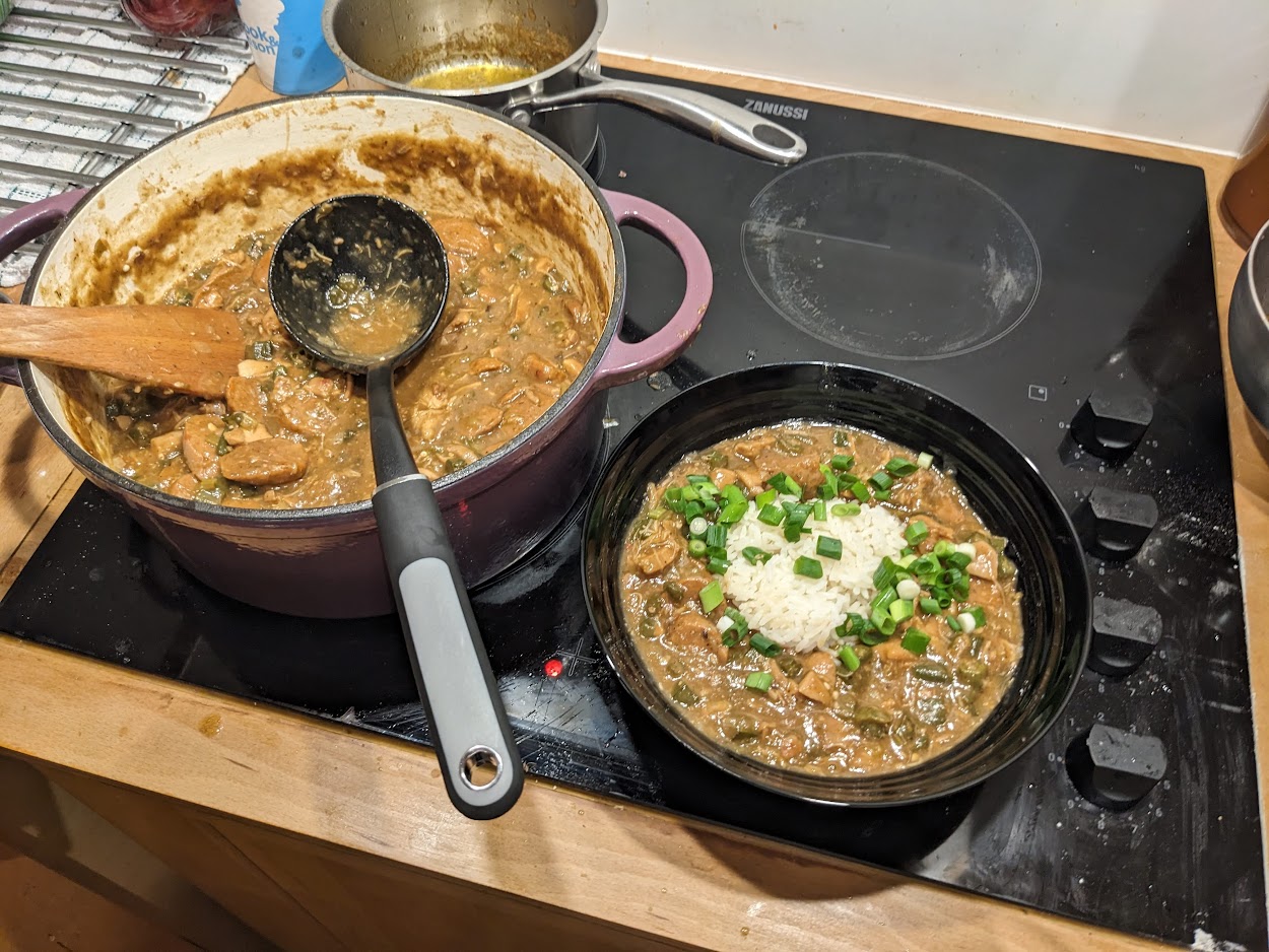 A large dutch oven full of gumbo on an electric stovetop. Inside the pot is okra, chicken, sausage, a ladle and a wooden spatula. Next to pot is a black bowl with rice and gumbo, topped with scallions.