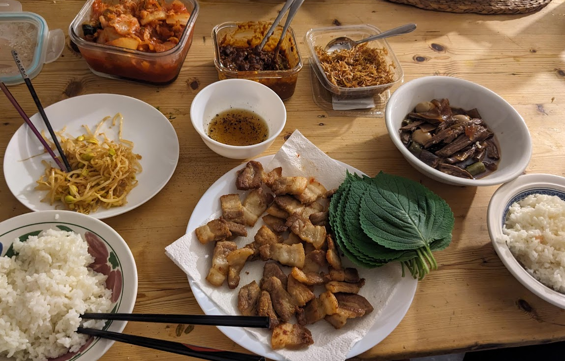 A table with rice, pork belly, perilla leaves, soy marinated beef (jang jorim), sesame dipping sauce, marinated bean sprouts, stir fried anchovies, ssamjang and kimchi. 