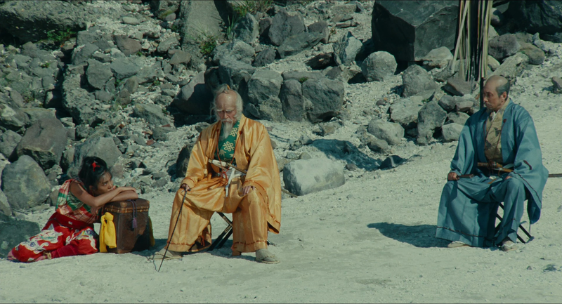 Hidetoro wears a yellow gown and sits in contemplation. The fool sits on the ground to his right and looks up at him. His advisor Tango wearing blue is sitting to his right several feet away. The background is rocky while the foreground is filled with white sand. The light in the frame is intense and unforgiving.